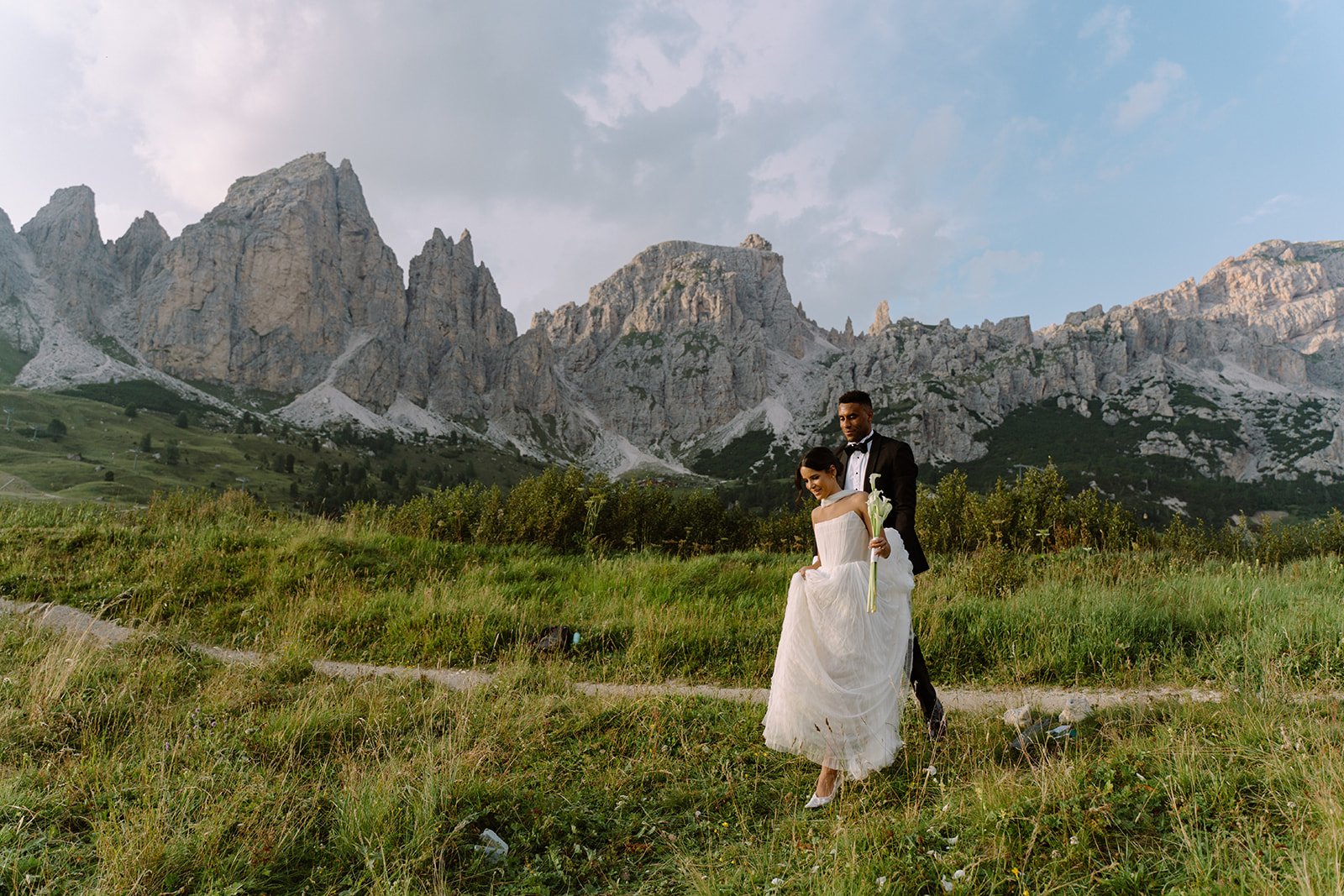 A bride and groom standing on a grassy field with mountains in the background. The bride is holding a white flower and wearing a white wedding dress; the groom is dressed in a black tuxedo.