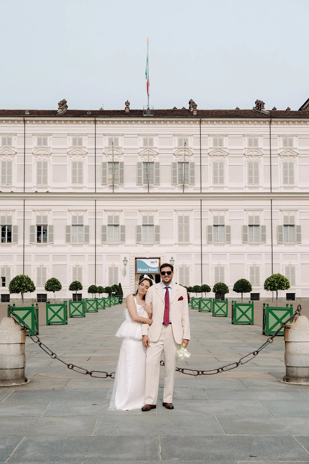 A couple dressed in wedding attire standing in front of a historic building with multiple windows and a flagpole on the roof. The woman is in a white wedding dress, leaning on the man, who is in a cream-colored suit with a red tie, holding a bouquet 