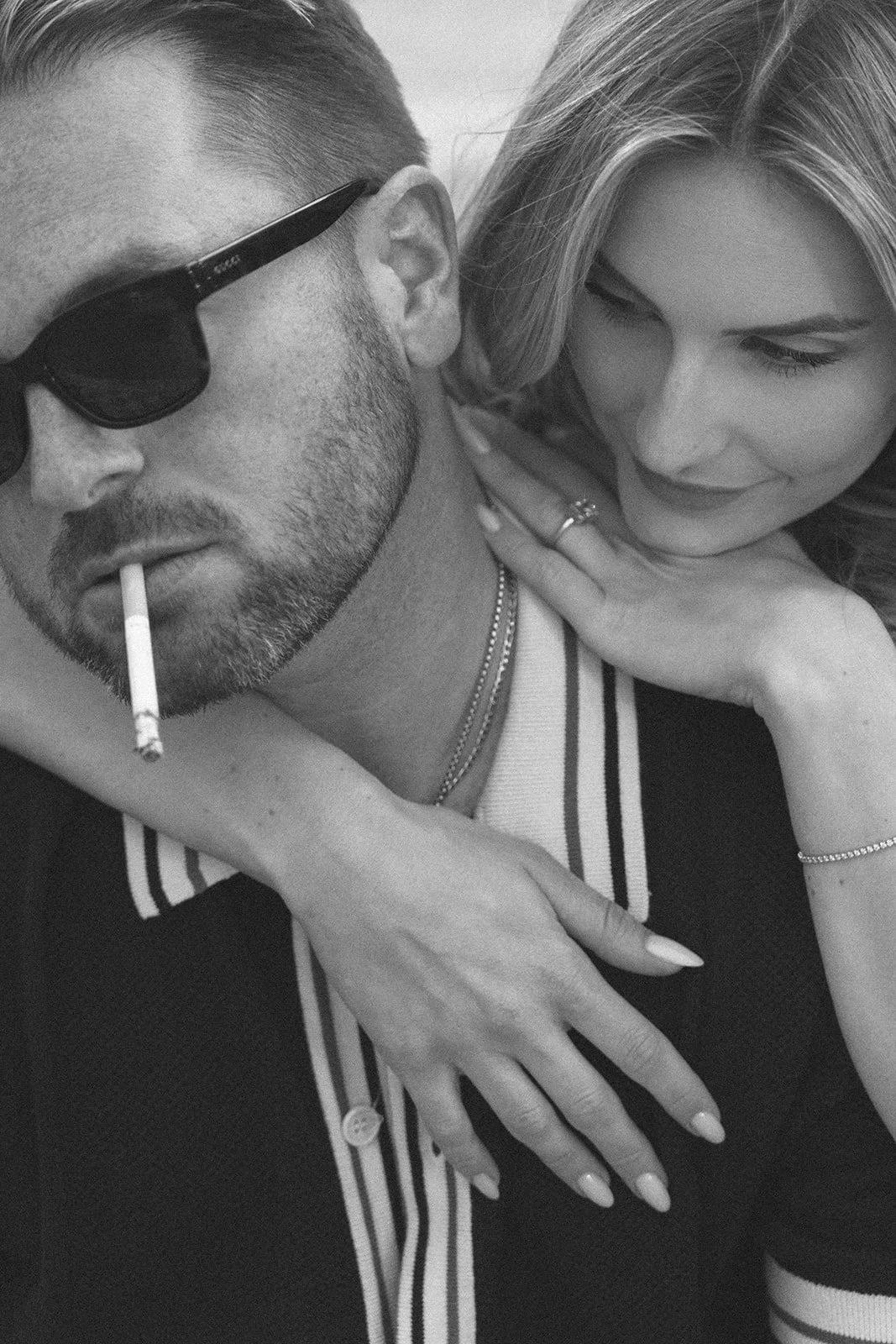 A black-and-white photo of a man wearing sunglasses smoking a cigarette, with a woman close beside him, both appearing relaxed and intimate.