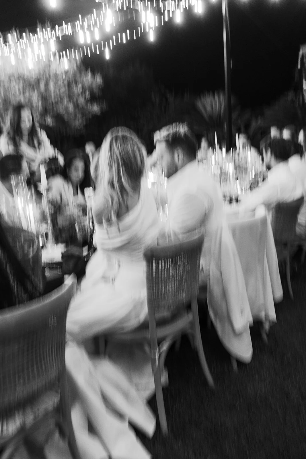 People sitting at a long table outdoors at night, surrounded by string lights, with bottles and glasses on the table.