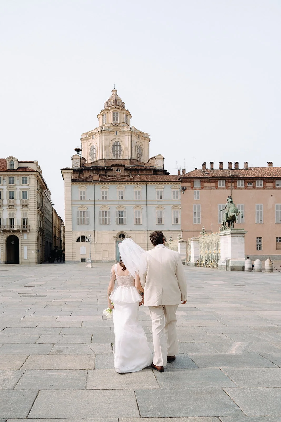 A bride and groom in wedding attire walking hand in hand in a city square with historic buildings and a statue in the background.