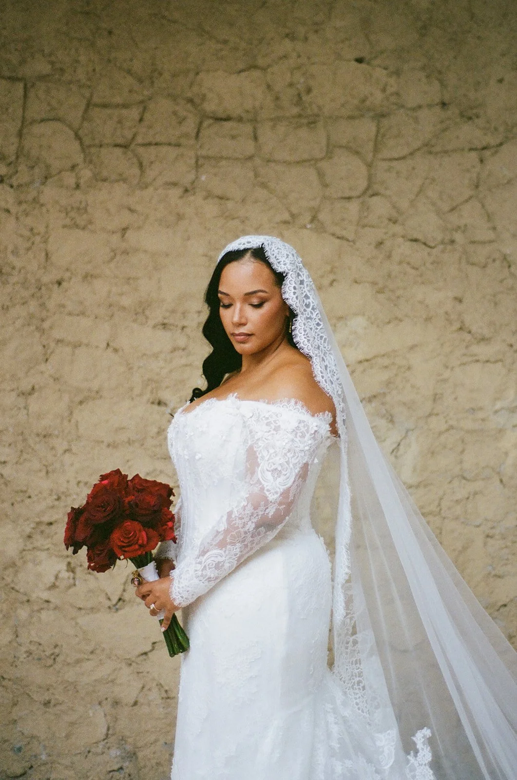 A bride in a white lace wedding dress with a lace veil, holding a bouquet of red roses, standing against a textured beige wall.
