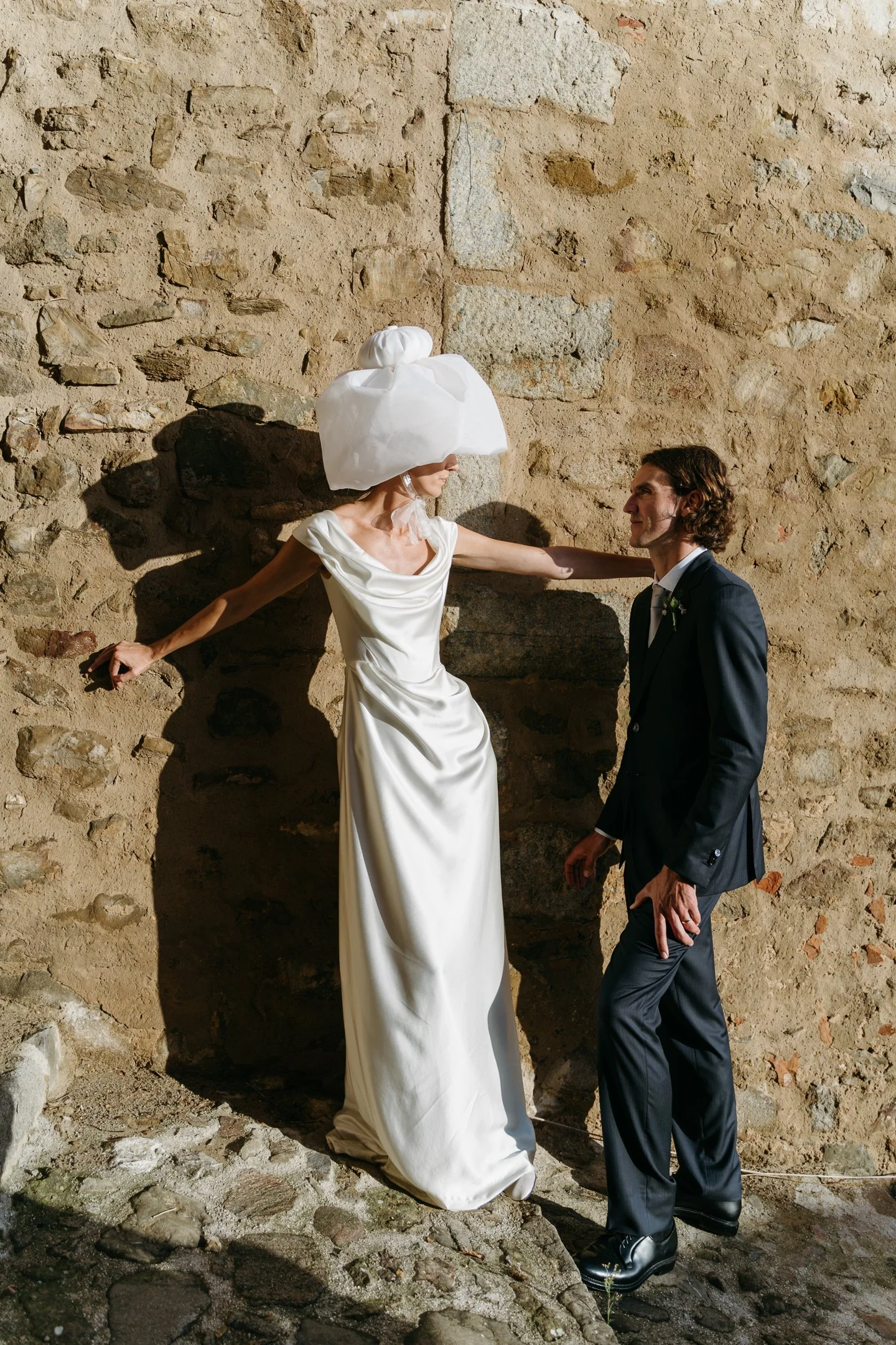 A woman dressed in a white wedding gown and elaborate headpiece leaning against a stone wall, looking at a man in a black suit and tie, standing near her during a wedding photo shoot.