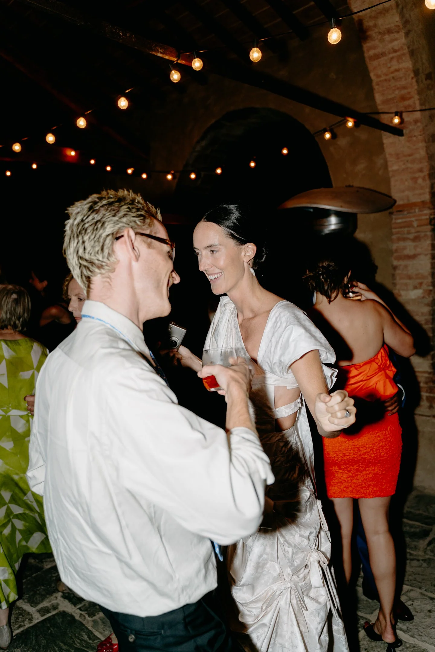Two people dancing and talking at a party in a dimly lit venue with string lights overhead. Other guests are visible in the background.