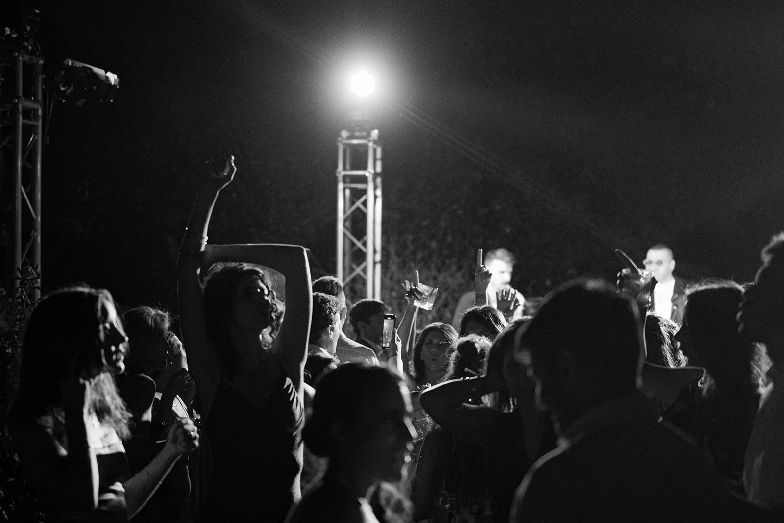 A black and white photograph of a crowded outdoor concert at night, with people dancing and listening to performers on stage. The stage is lit by a bright overhead light, and the audience members are raising hands and taking photos.