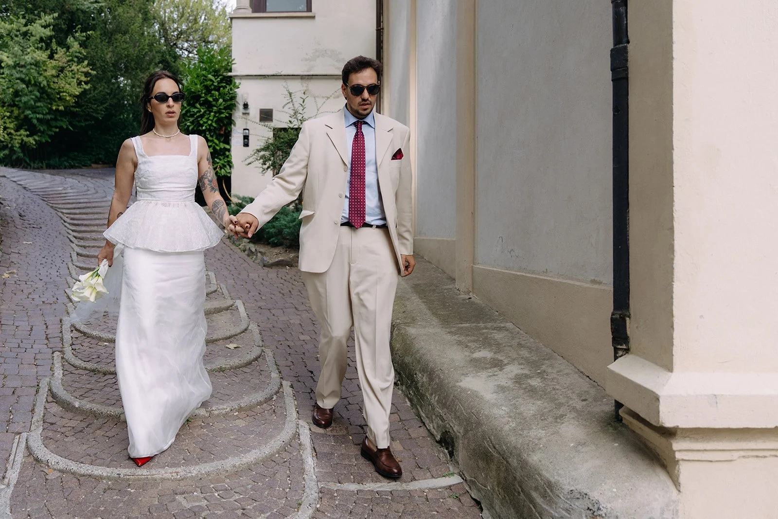 A man and a woman holding hands and walking along a cobblestone path, with the woman holding a small bouquet of white flowers. They are dressed in formal attire, with the woman in a white gown and the man in a light-colored suit, both wearing sunglas