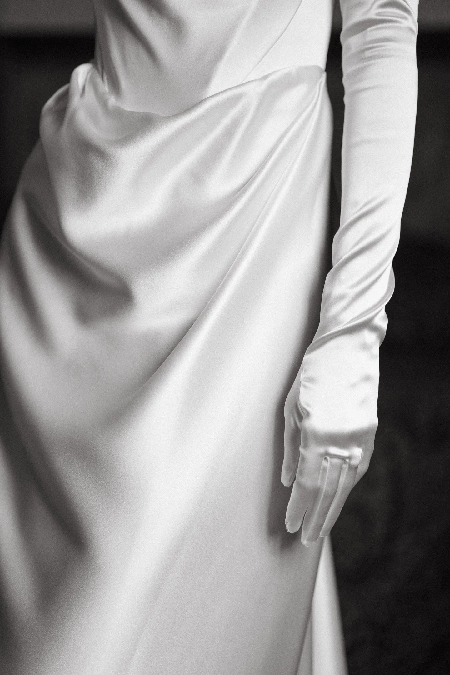 Close-up of a person wearing a white satin long-sleeve dress and matching white satin glove, with a dark background.