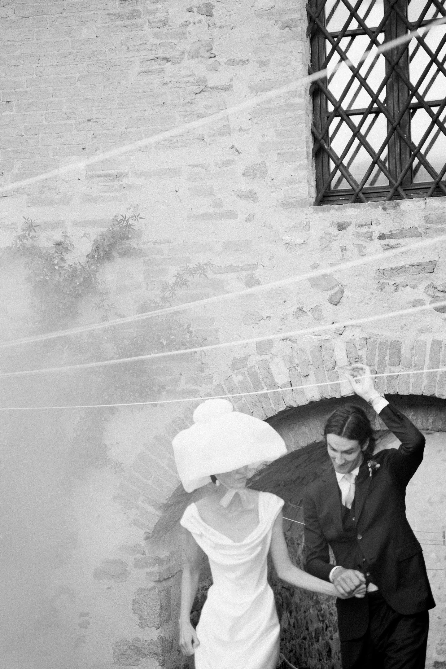 Black and white photo of a man and woman walking hand in hand under an arched brick bridge, with laundry lines overhead and a window with bars on the brick wall.
