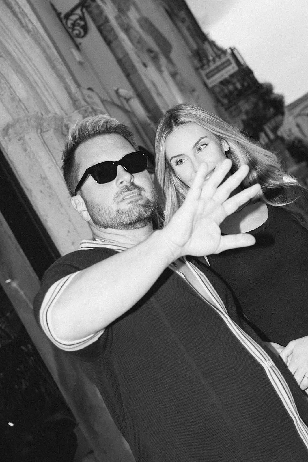 A man wearing sunglasses and a woman standing behind him, both outdoors on a building sidewalk with ornate architecture in the background. The man is reaching out with his hand.