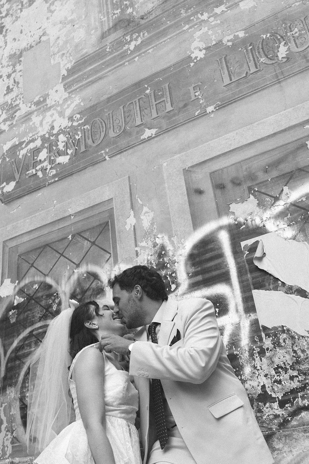 Black and white photo of a couple in wedding attire sharing a kiss in front of a wall with a sign reading 'Vermouth & Licores'. The wall has peeling paint, and there is graffiti art overlaying the photo of the couple.