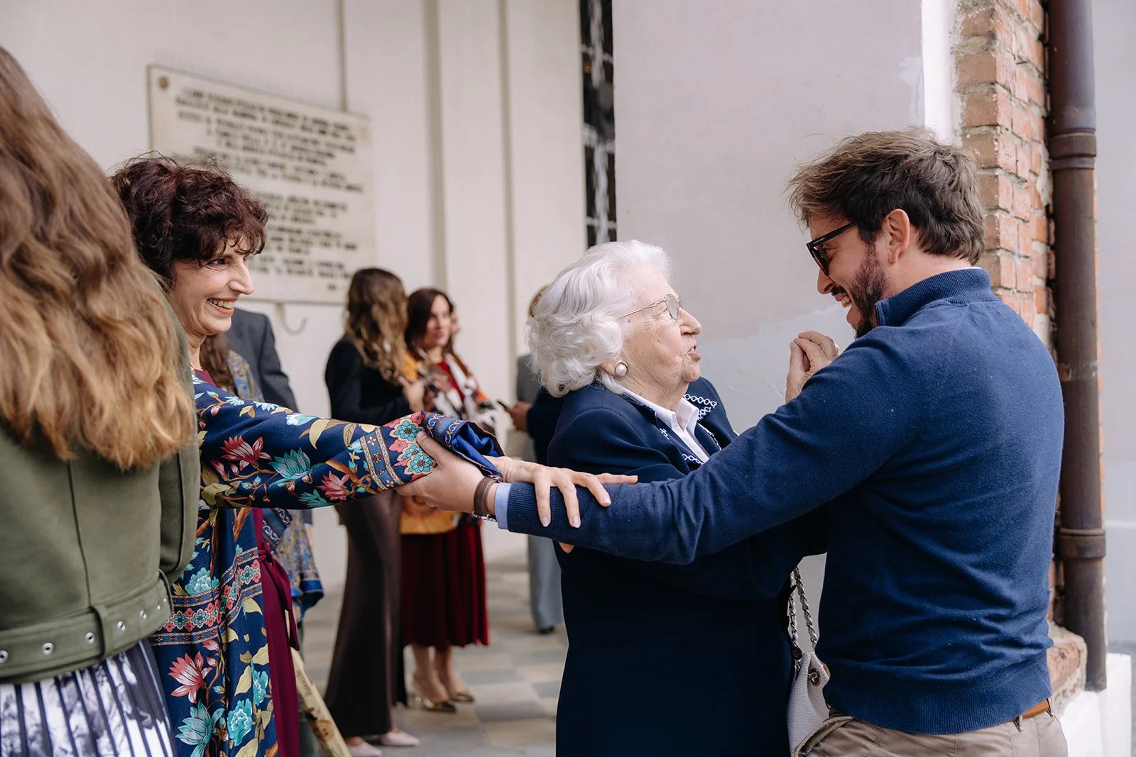 A group of people holding hands in a line, smiling and interacting with an elderly woman in a dark blazer. The setting appears to be an event or gathering near a brick wall.