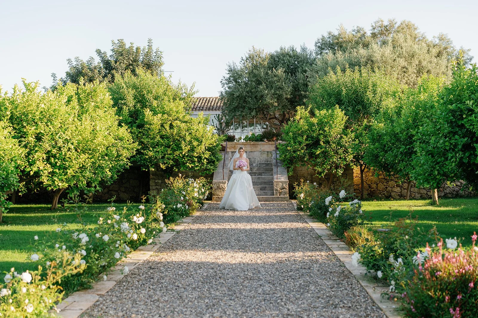 A bride in a wedding dress holding a bouquet of pink flowers, standing on stone steps in a lush garden with green trees and flowering plants.