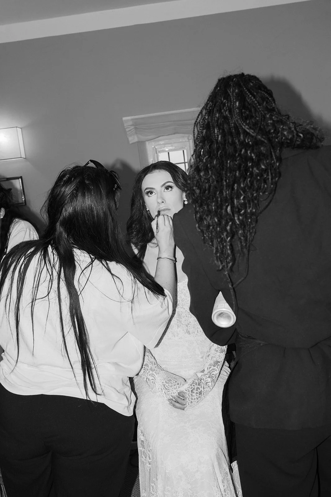 A woman in a wedding dress getting makeup applied by a stylist, surrounded by three other women, in a room with a small window and wall-mounted light.