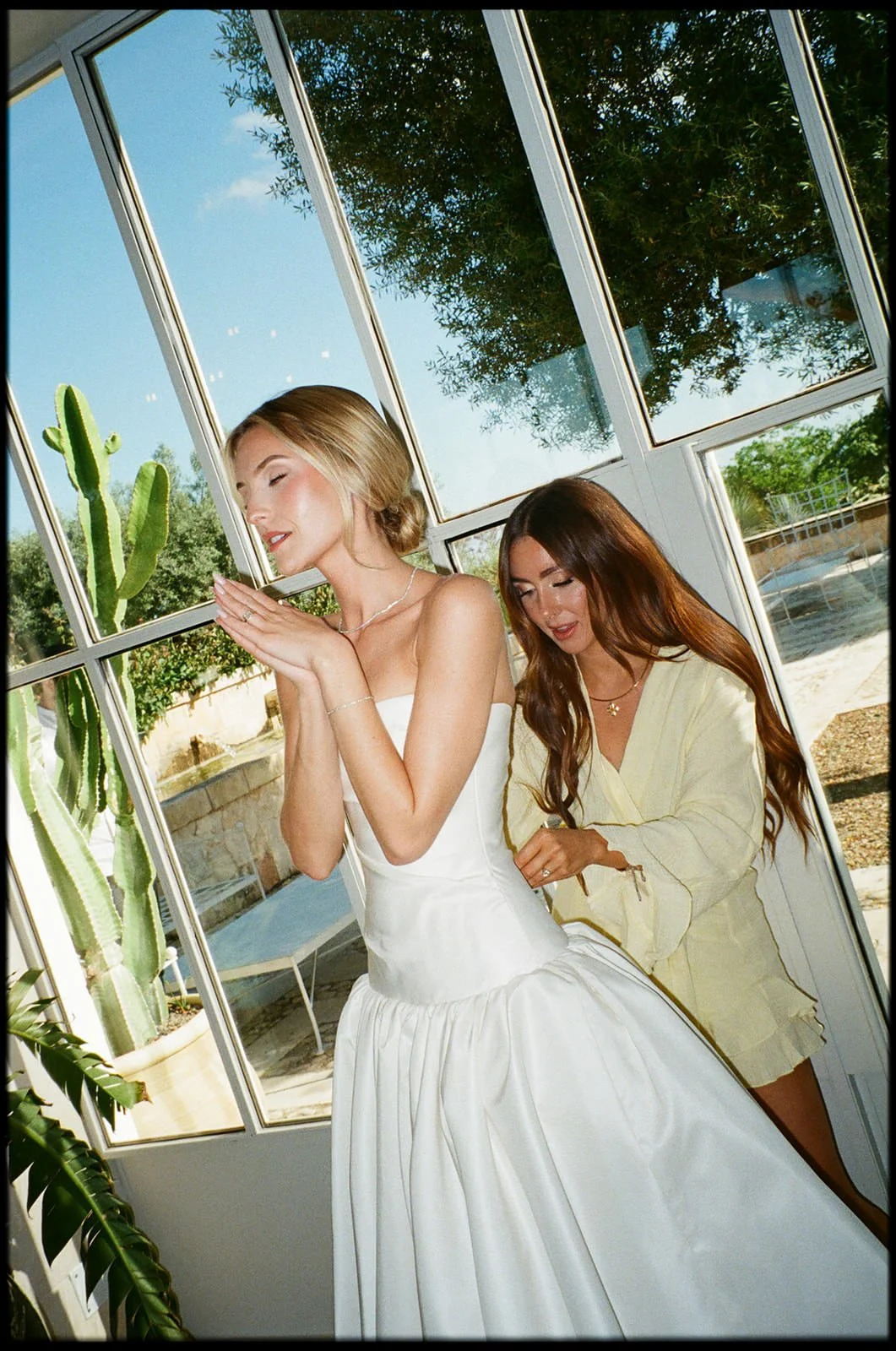 Two women inside a bright room with large windows and greenery outside. One woman is wearing a white dress, and the other is in a yellow outfit. The woman in white appears to be posing with closed eyes and hands together, while the other is helping h