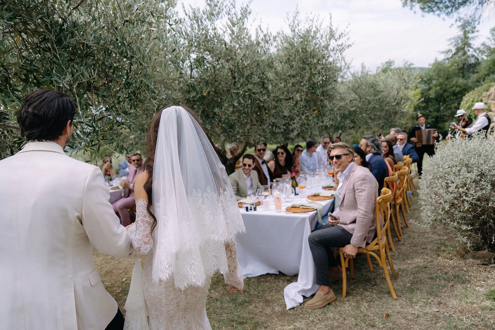 Bride and groom standing outside at a wedding reception, facing a long table filled with guests, with trees and greenery in the background.