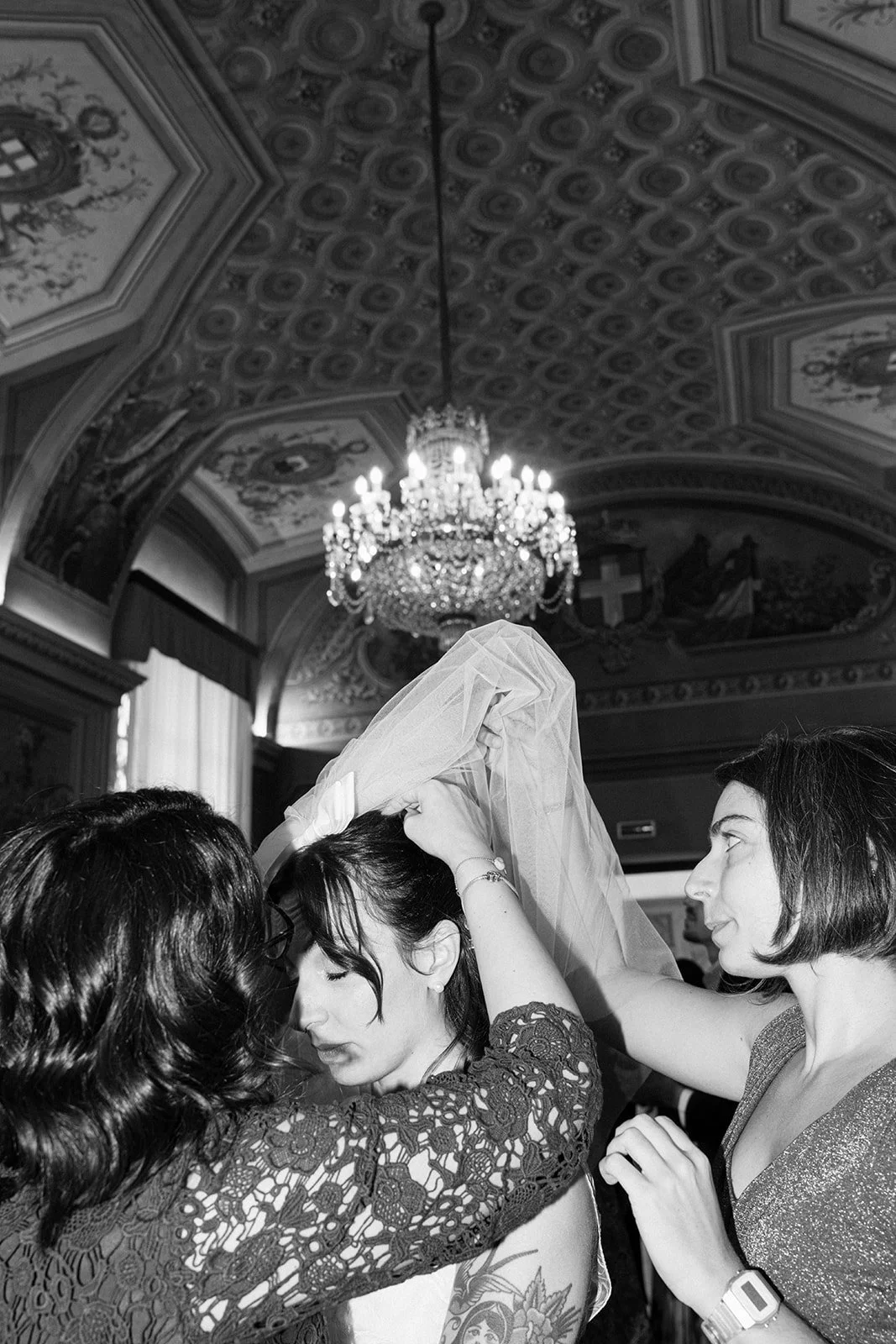 A woman in a lace dress has a veil placed on her head by two other women in a decorated room with a chandelier.