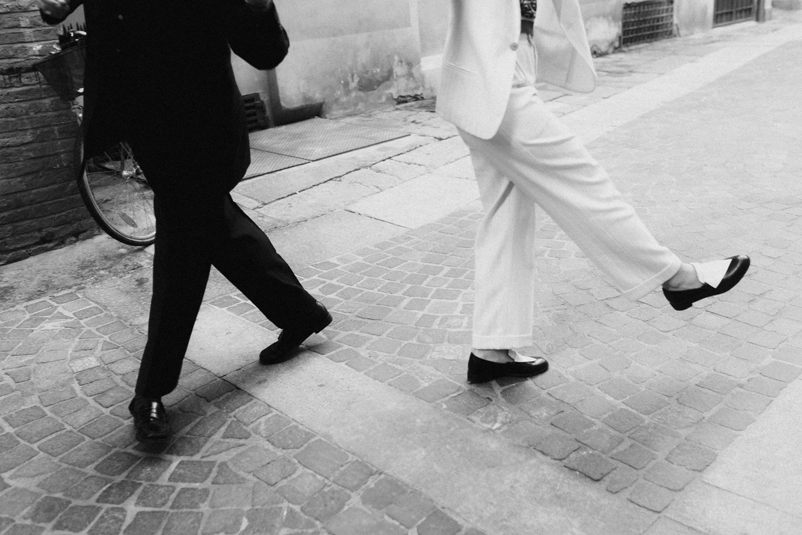 People walking on a cobblestone street, legs and feet visible, dressed in formal clothing; one in a black suit and the other in a white suit.