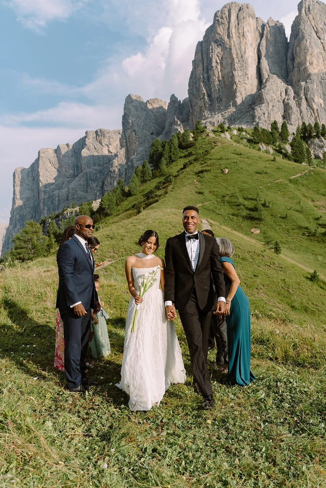A wedding ceremony outdoors on a grassy hillside with rocky mountains in the background, featuring a bride in a white dress and a groom in a black tuxedo holding hands and smiling.