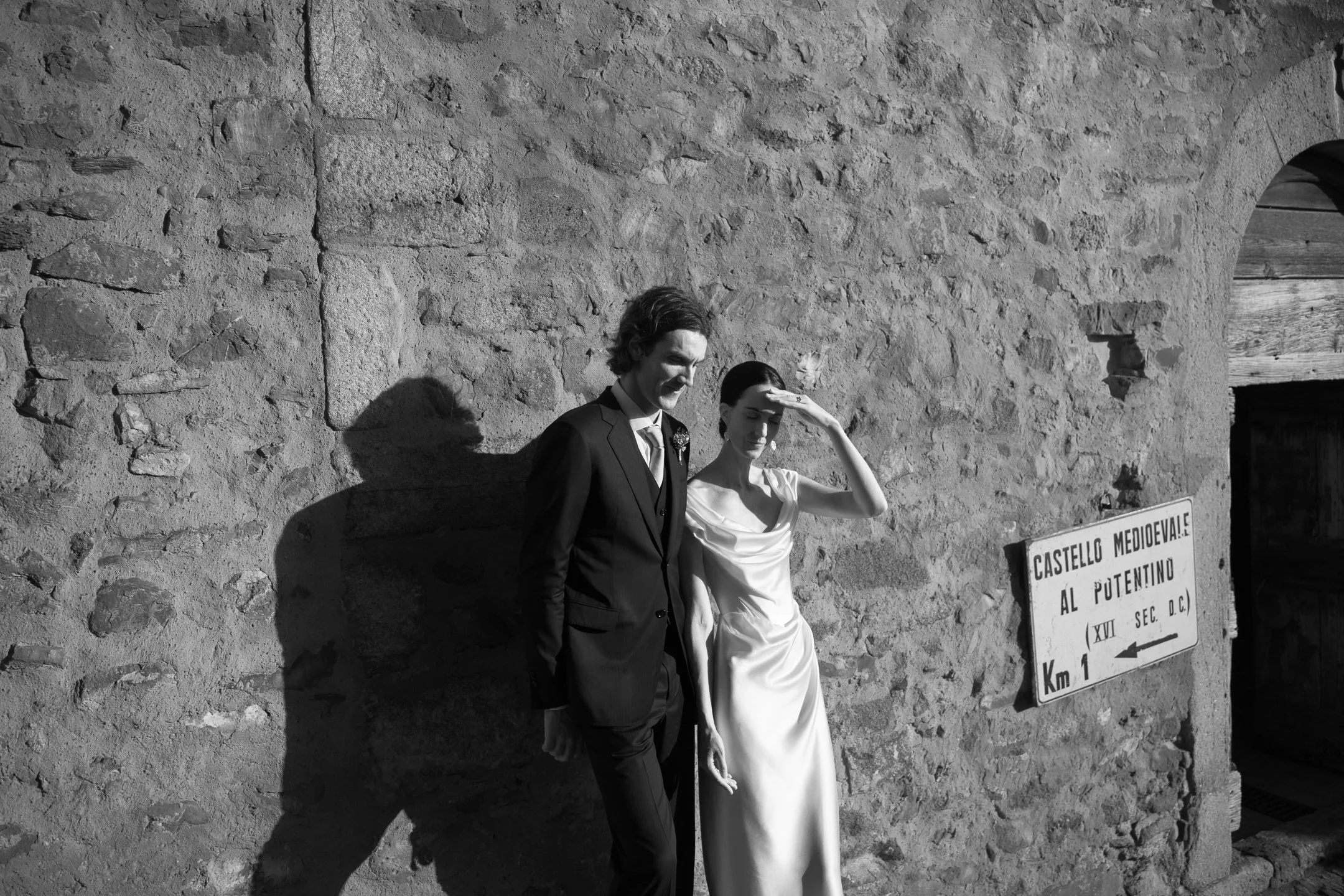 A black and white photo of a man and woman in wedding attire standing against a stone wall. The woman is shielding her eyes from the sun with her hand, and the man is smiling. There is a sign on the wall pointing to the medieval castle.