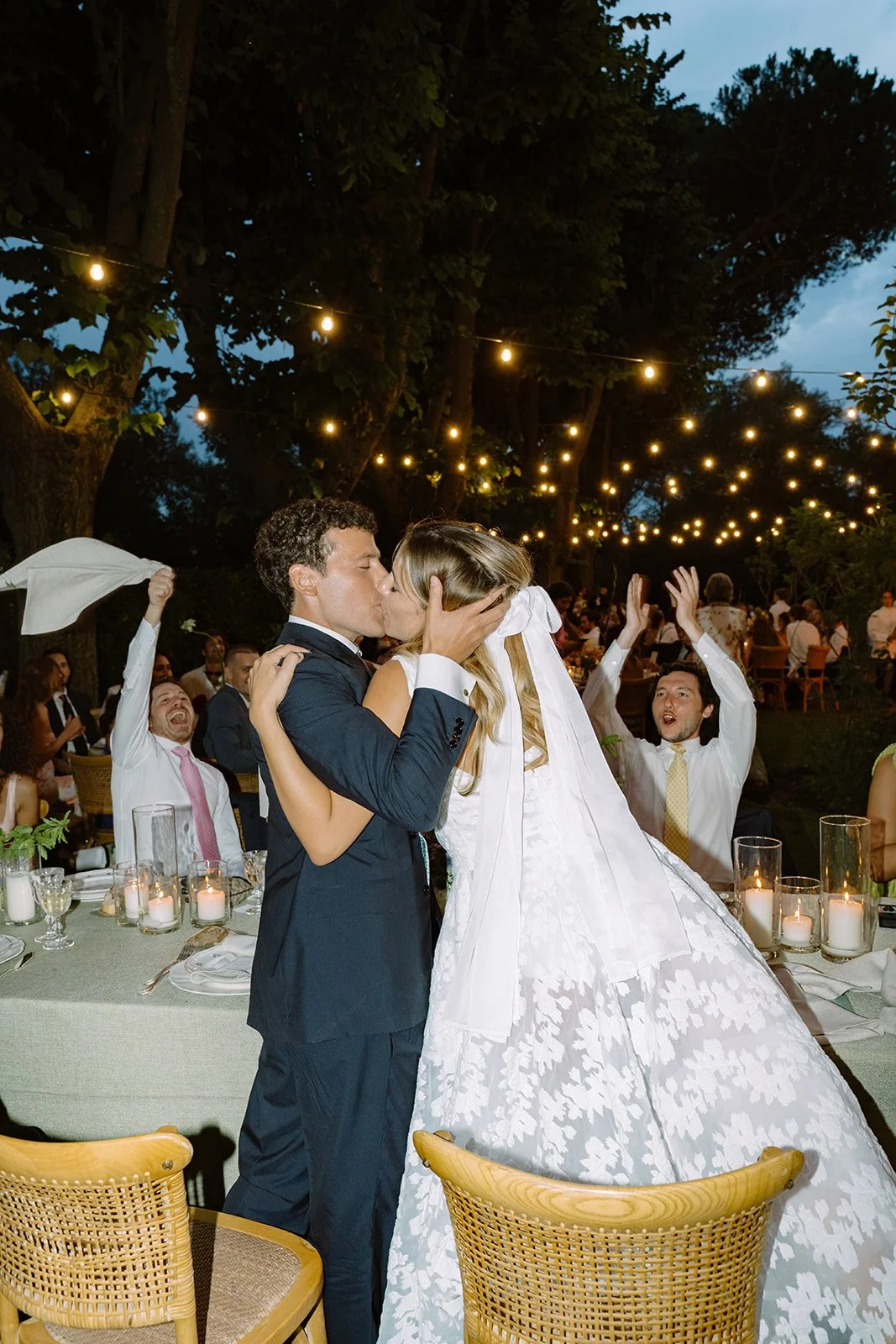 A wedding celebration outdoors at night, couple sharing a kiss, surrounded by guests clapping and celebrating under string lights.