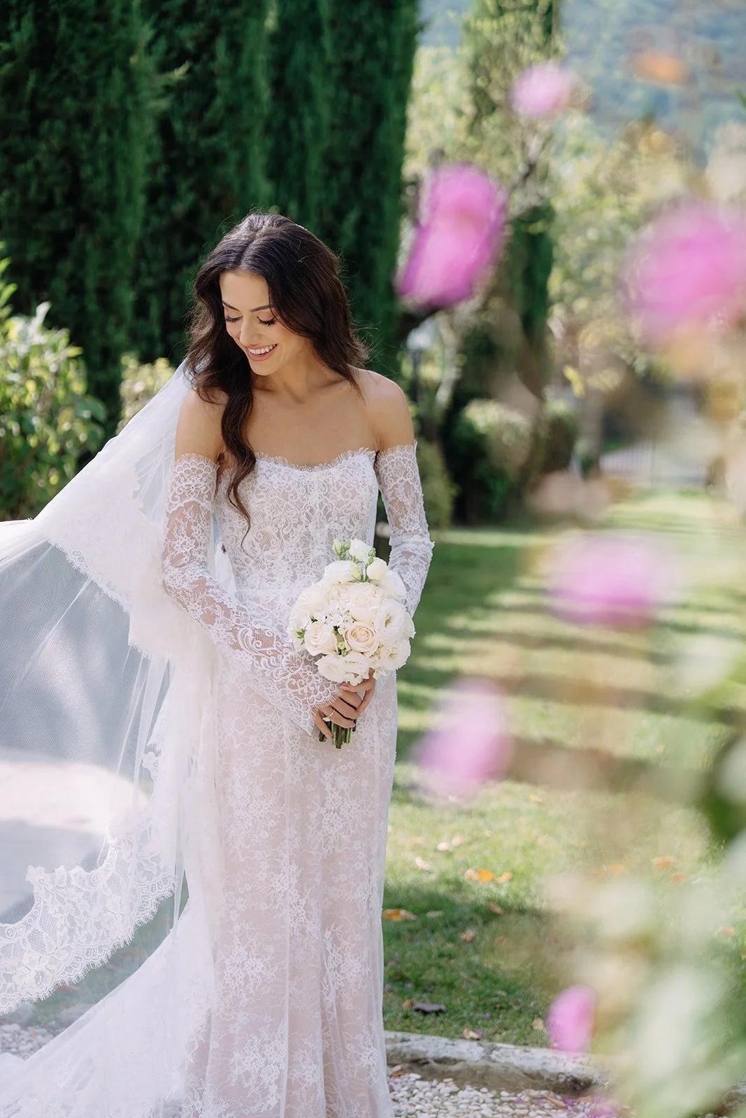 A bride in a lace wedding dress holding a bouquet of white roses, smiling outdoors in a garden setting with trees and pink flowers in the foreground.