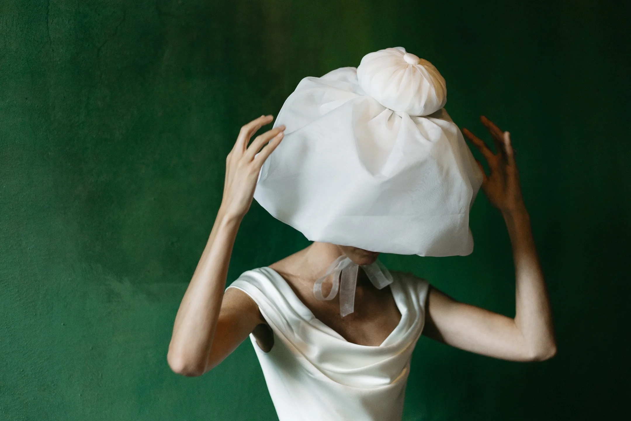 Woman with a large white fabric wrapped on her head, standing against a green background, wearing a cream satin dress.