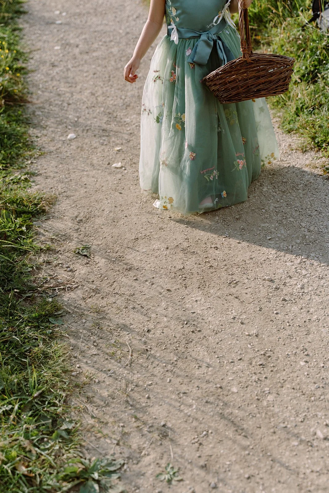 Person walking on a dirt path, wearing a long, embroidered green dress with a sash and carrying a wicker basket.
