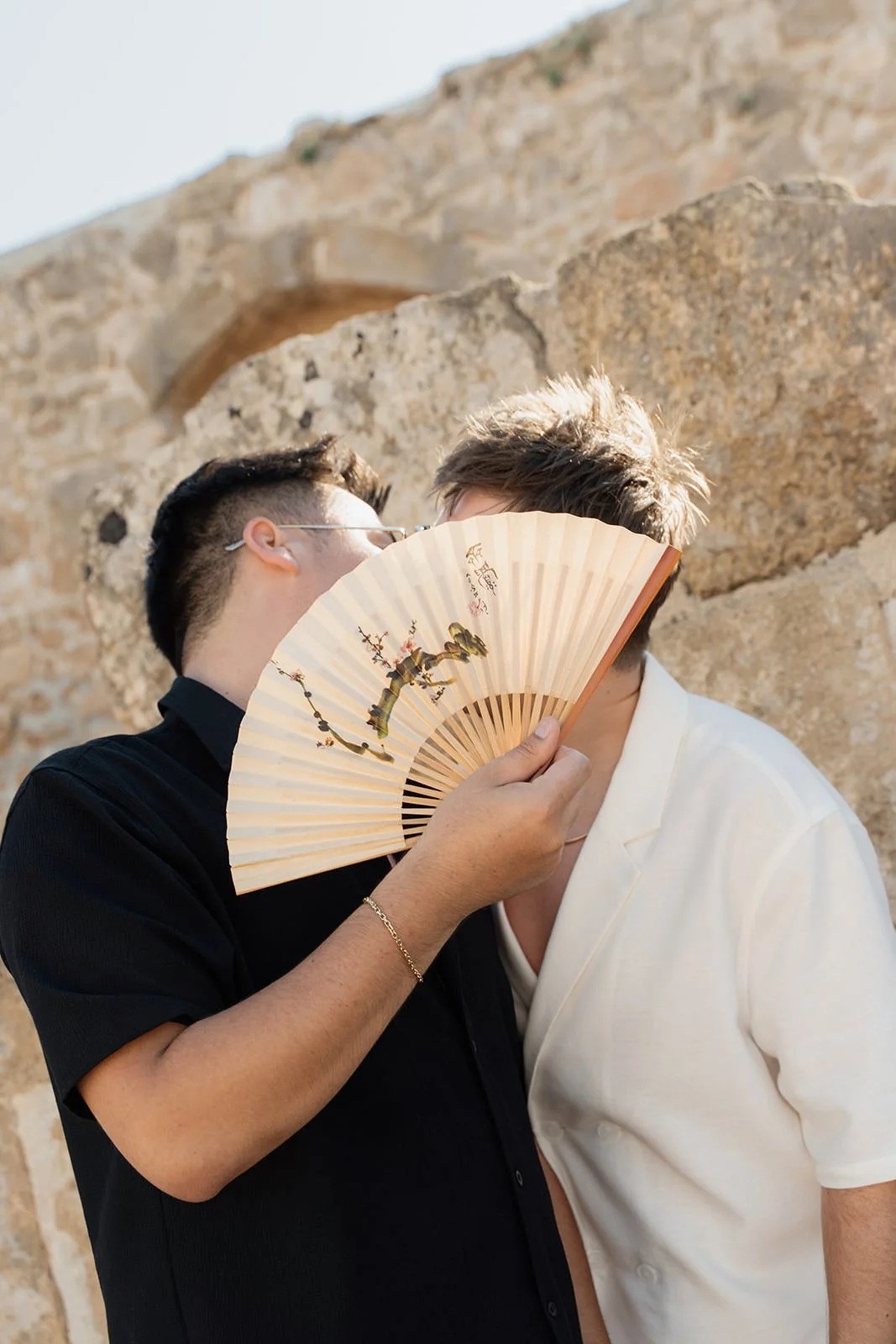 Two men standing close together behind a fan, with a stone wall in the background.