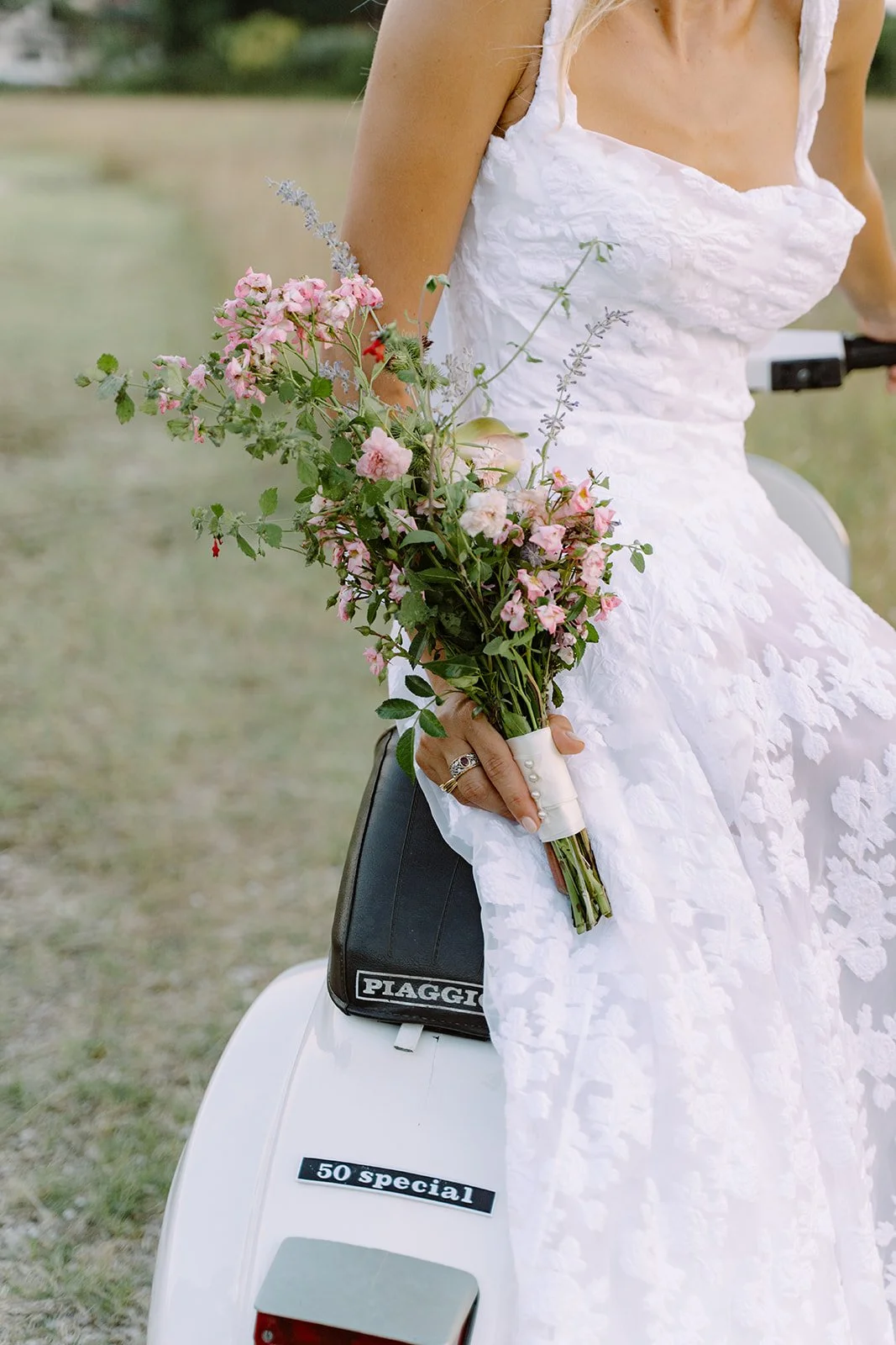 A woman in a white dress holding a bouquet of pink and purple flowers, sitting on a white scooter with a black bag that has 'PIAGGIO' written on it and a sticker that reads '50 special'.