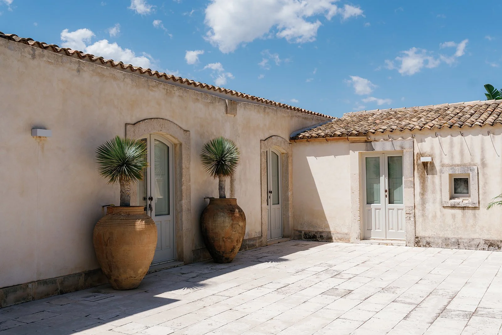 A courtyard with light-colored stone tiles, beige stucco walls, and terracotta roof tiles. There are two doors with white frames and glass panels, flanked by two tall potted plants with spiky green leaves in large clay pots. The sky is blue with scat
