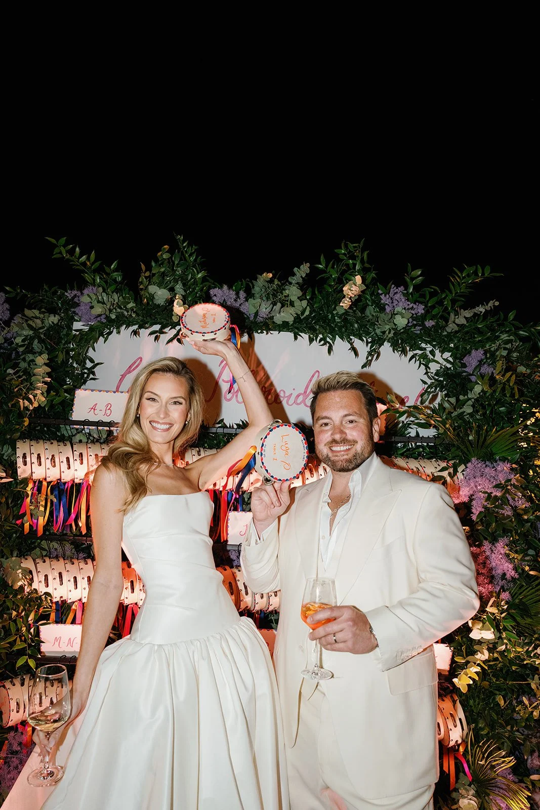 A smiling woman in a white strapless dress holding a small birthday cake, and a man in a white suit light pink shirt holding a drink and a small polka-dotted cake, standing in front of a decorative backdrop with green foliage, pink flowers, and color