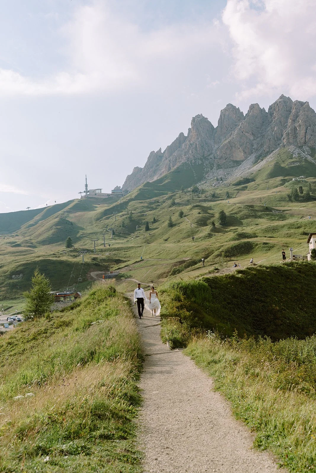 A bride and groom walking hand-in-hand along a narrow dirt trail in a lush green mountain landscape under a partly cloudy sky.