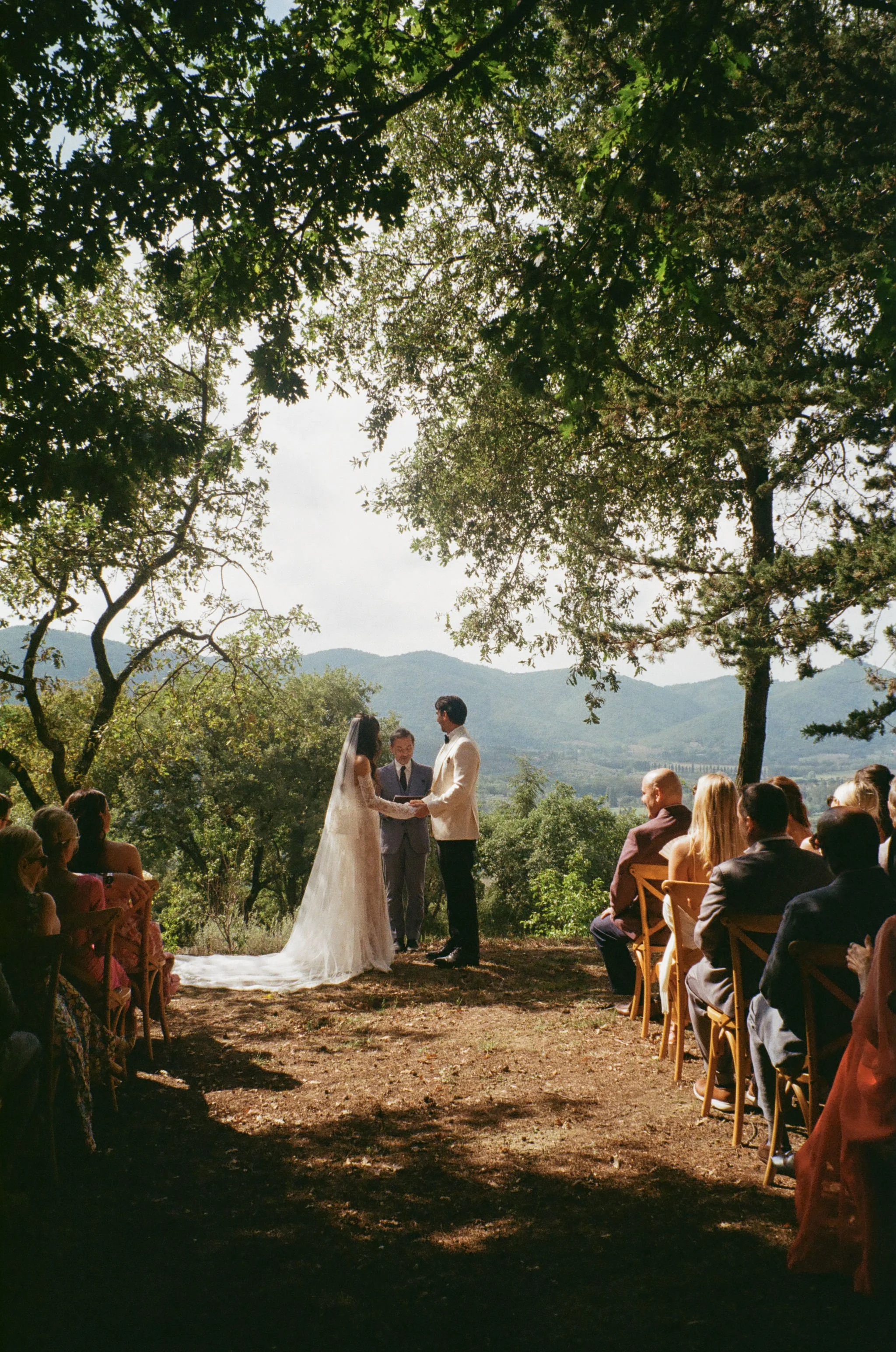 A couple getting married in an outdoor ceremony under trees, with guests seated on wooden chairs.