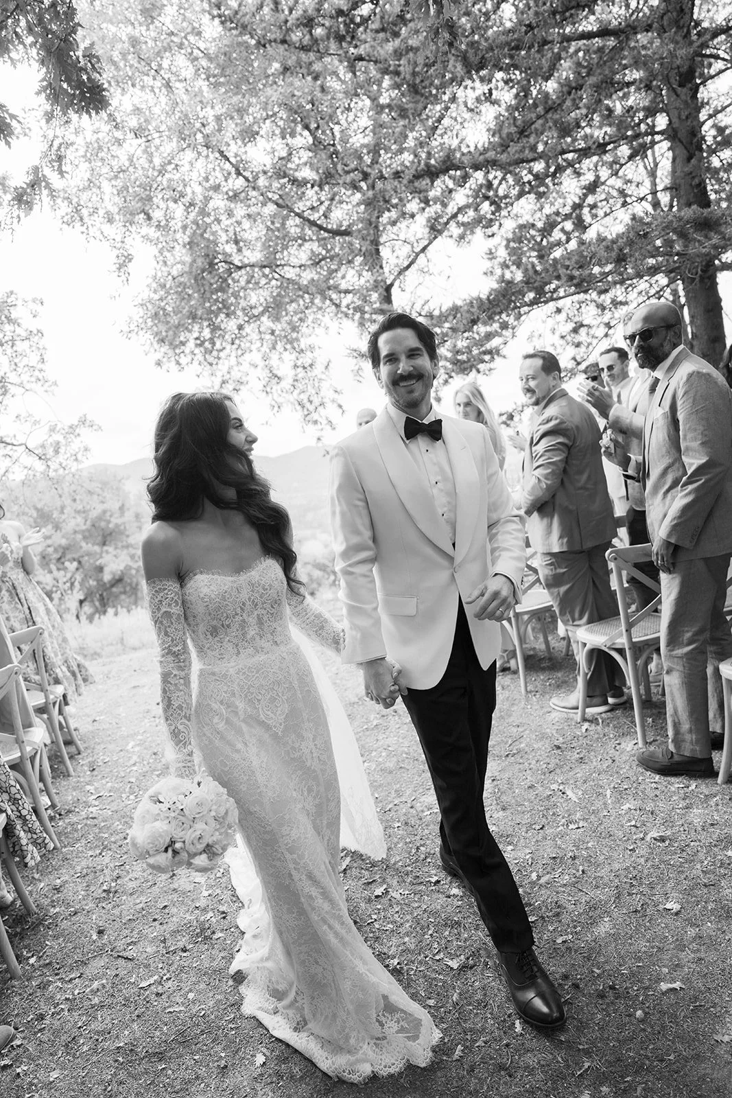 Black and white photo of a bride and groom walking hand in hand outdoors, surrounded by wedding guests, with trees overhead.