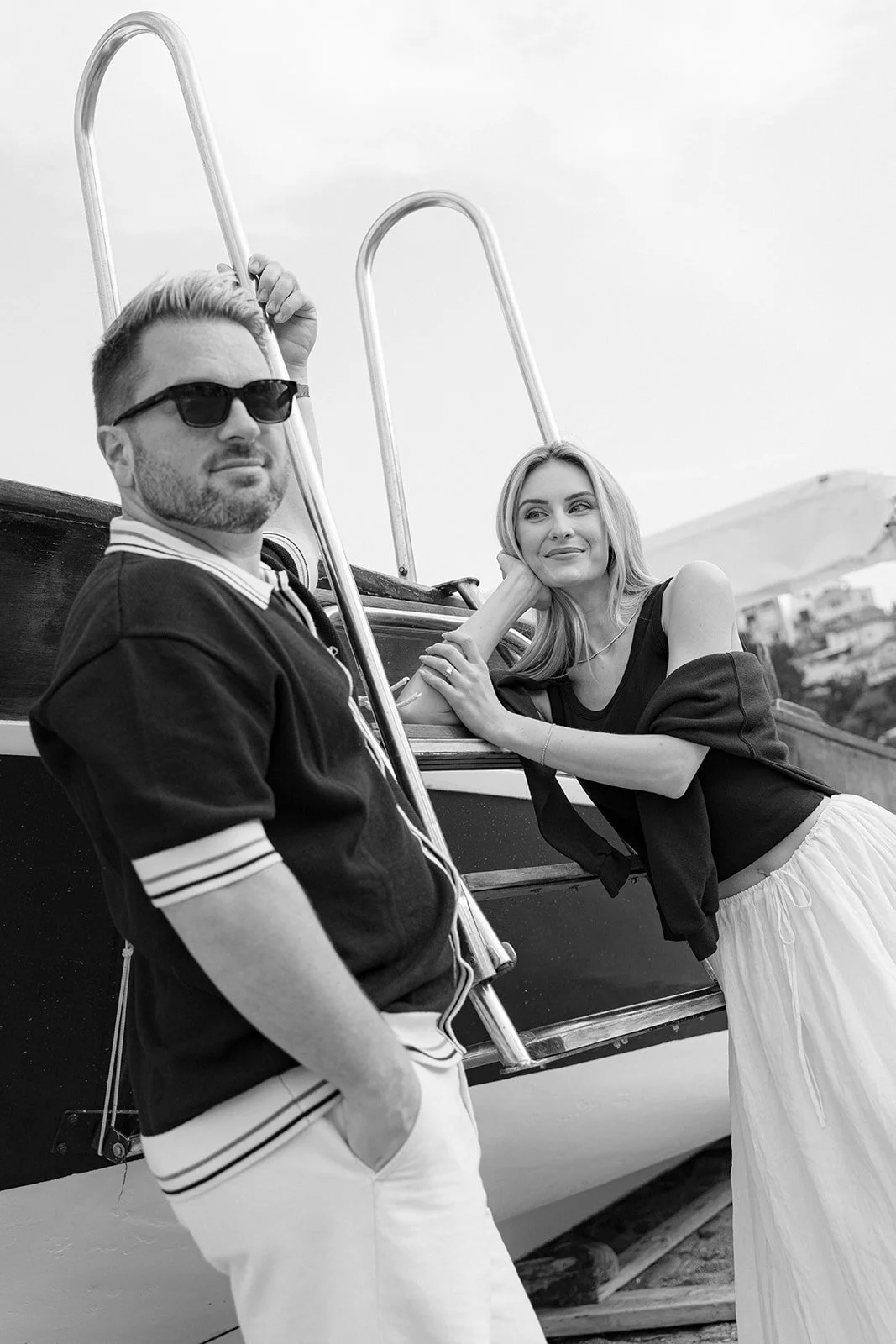 A black-and-white photo of a man with sunglasses and a woman with long hair, posing near a boat, with a hillside and buildings in the background.