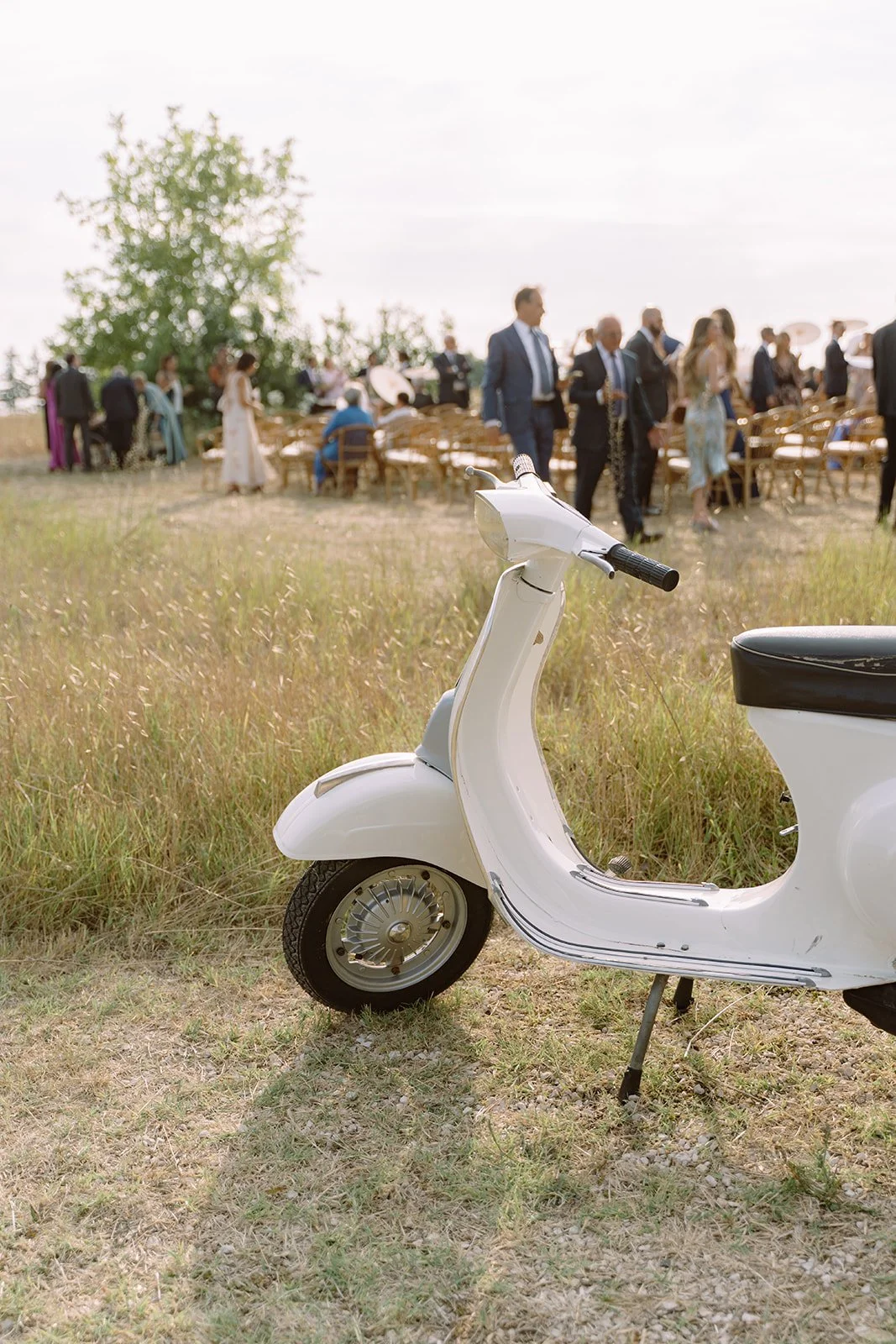 White vintage scooter parked on grass at an outdoor wedding ceremony with guests and chairs in the background.