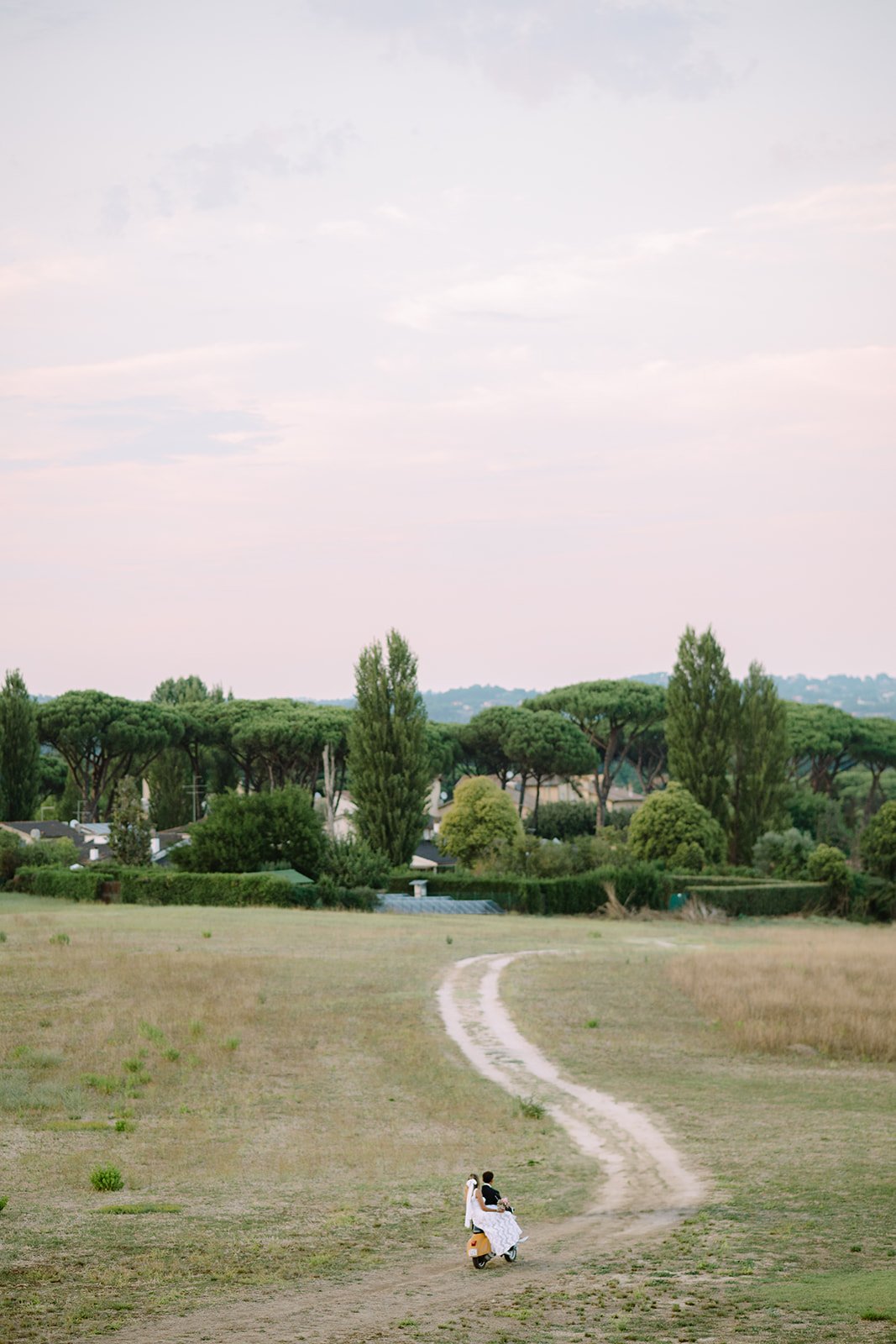 A couple riding a scooter along a dirt path in a grassy field with trees in the background under a cloudy sky.