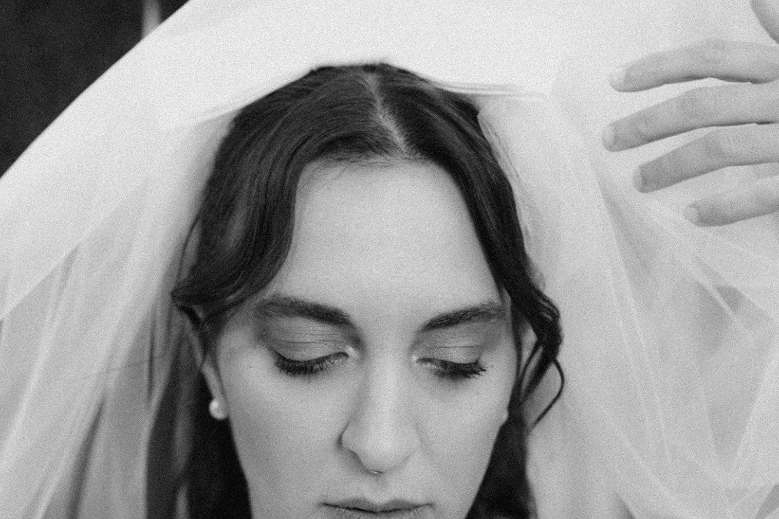 Black and white photo of a bride with closed eyes, dark hair, and pearl earrings, lying with a veil over her head.