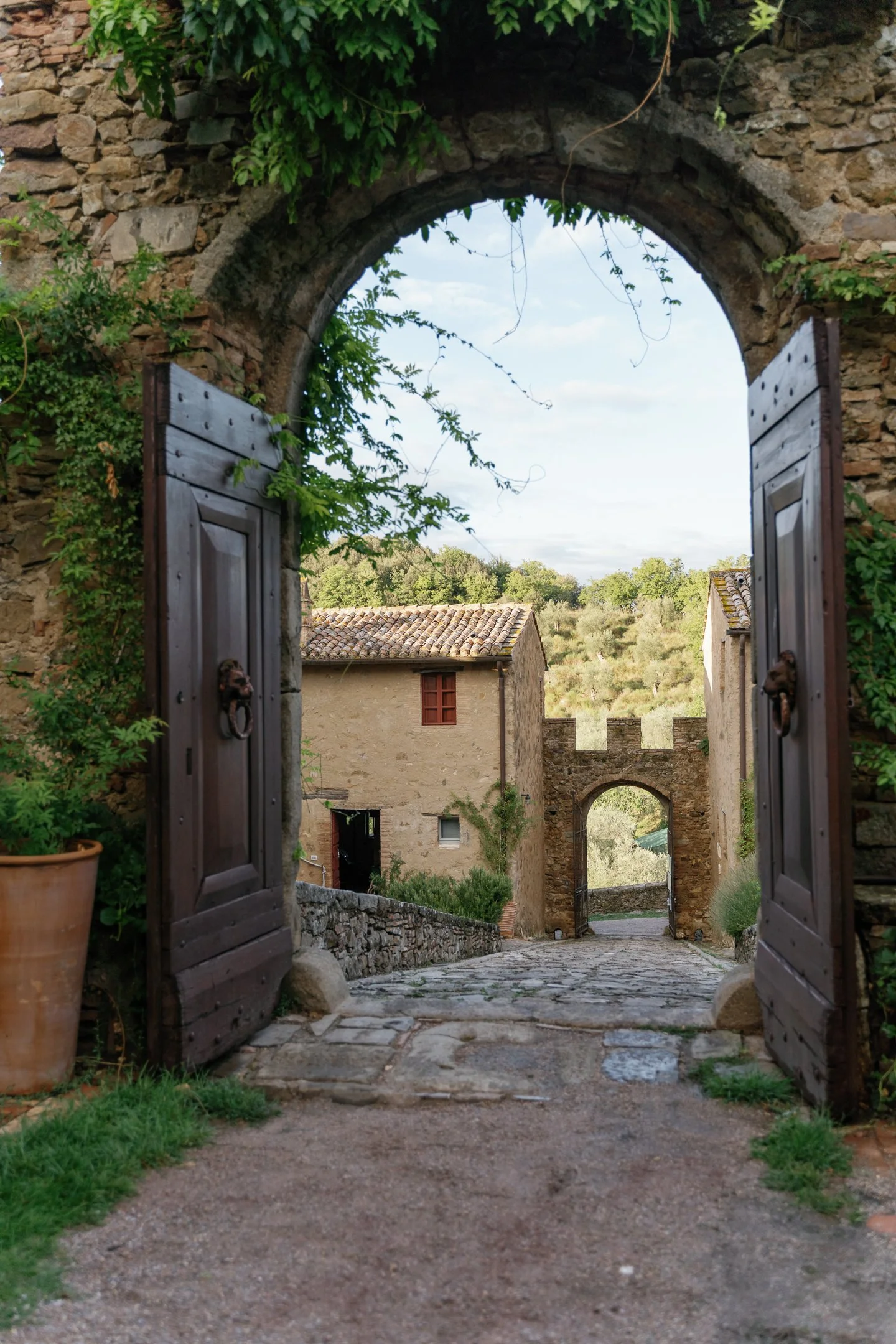 A scenic view through an arched stone doorway with open wooden shutters, showing a cobblestone path, rustic buildings, and green hills in the background.