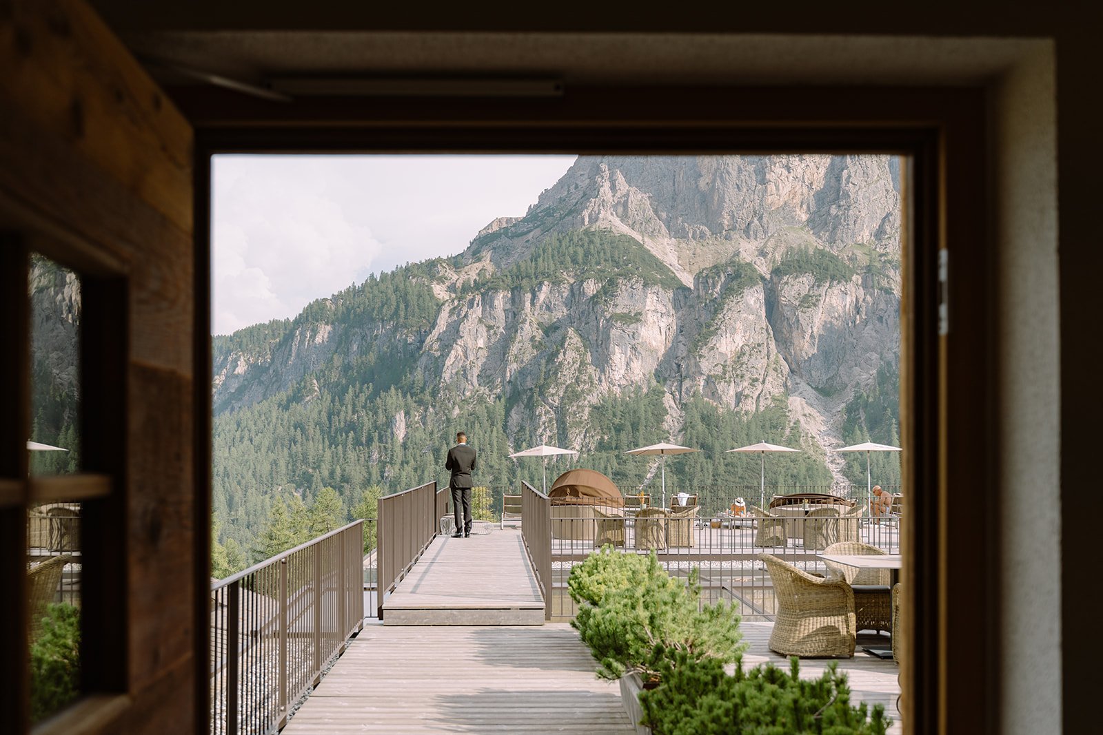 A mountain view framed by a window, showing a wooden deck with a person in a suit looking at the mountains, outdoor furniture, and umbrellas.