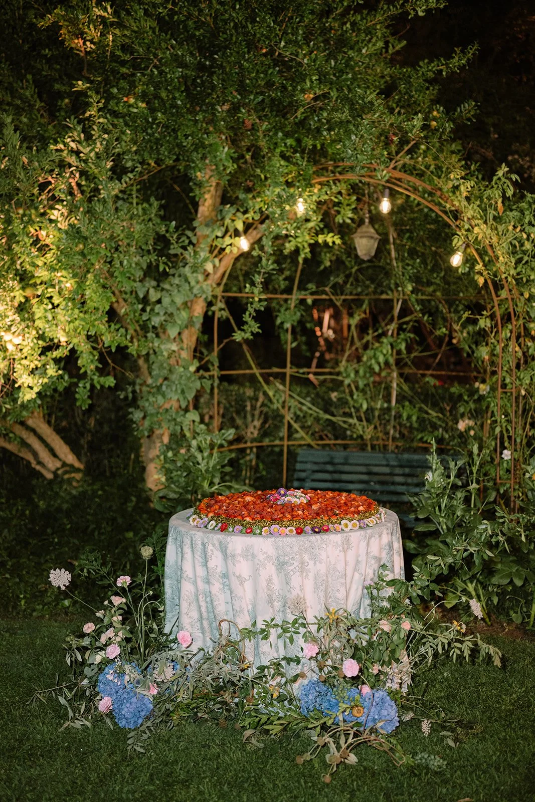 Outdoor celebration table with a floral arrangement on a white tablecloth, surrounded by greenery and string lights at night.