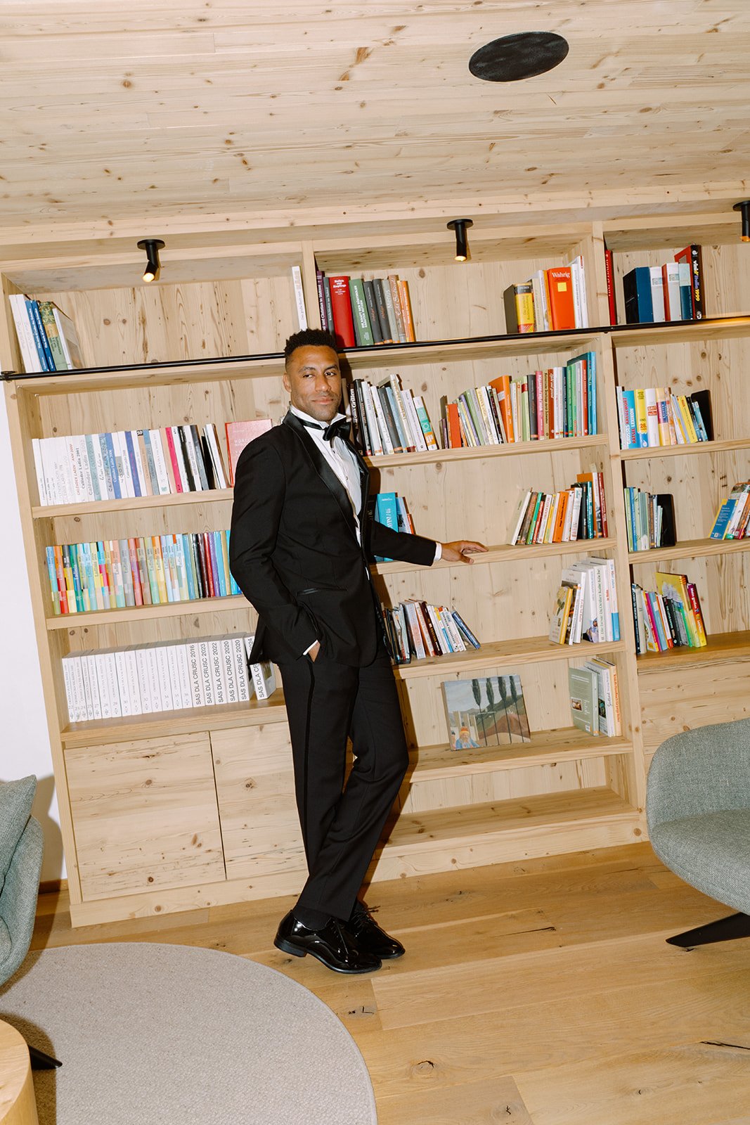A man in a tuxedo poses in front of wooden bookshelves filled with colorful books in a modern room with wooden flooring and ceiling. There are two light fixtures on the ceiling.