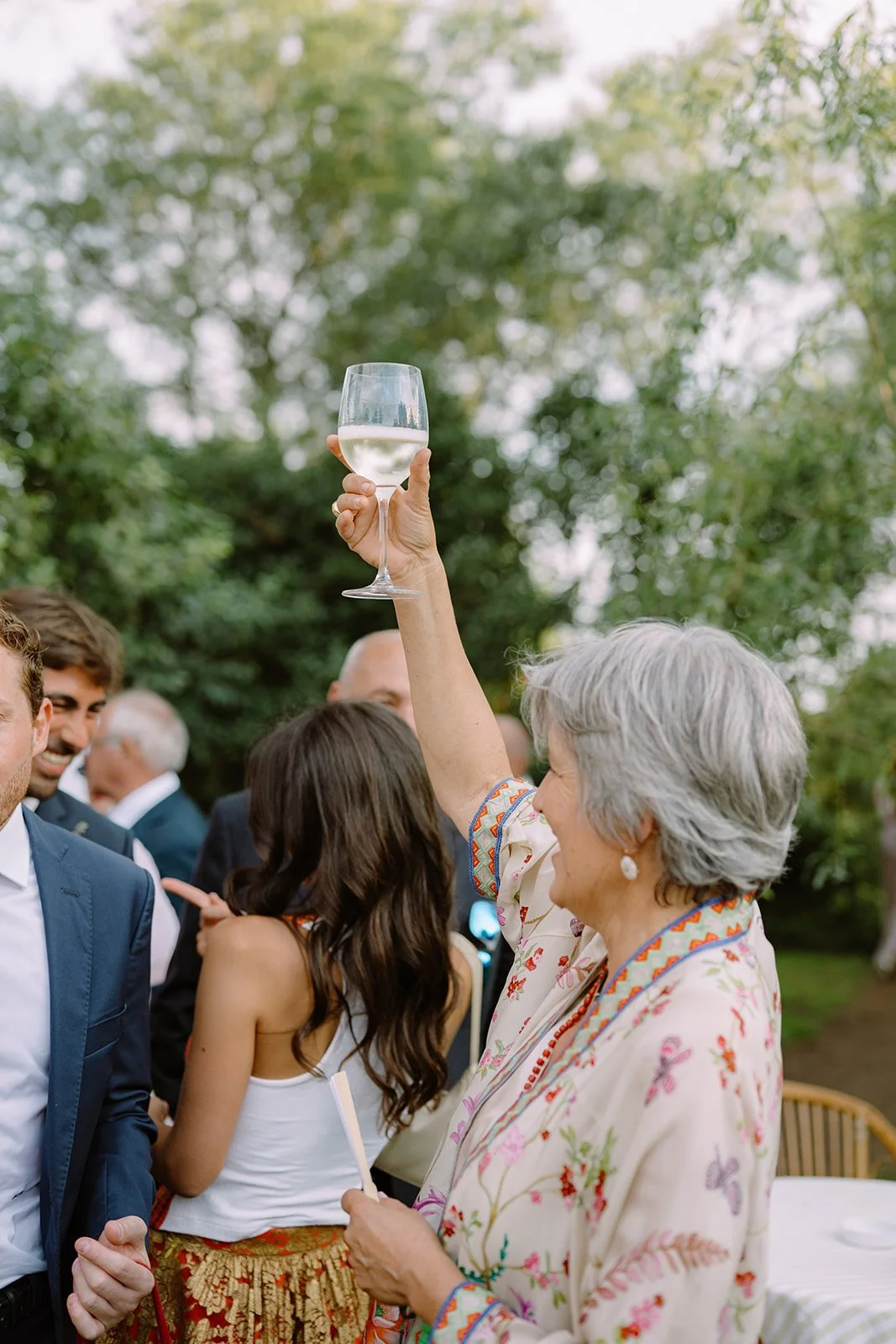An elderly woman with gray hair holding up a glass of white wine at an outdoor social gathering, surrounded by people in formal attire.