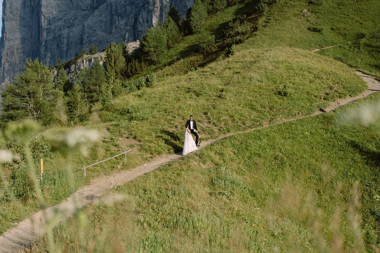 A bride and groom standing on a narrow dirt trail on a grassy hillside with trees and cliffs in the background.
