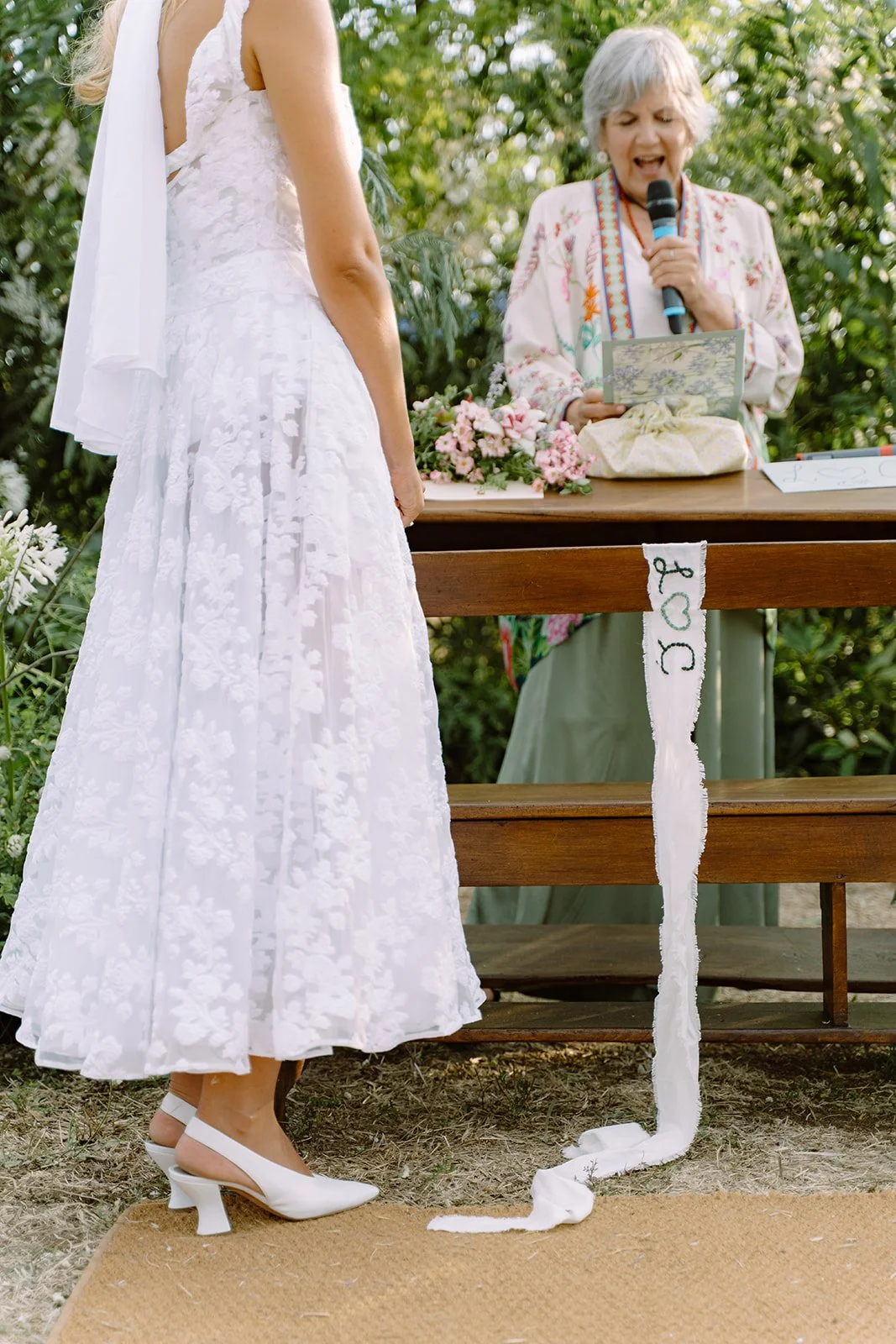A bride in a white lace dress and white heels standing in front of a woman with gray hair, reading vows at an outdoor wedding ceremony.