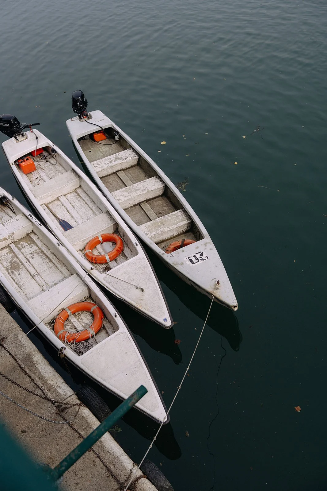 Three small white boats docked at a concrete pier on calm water, each with orange life rings inside and outboard motors attached.