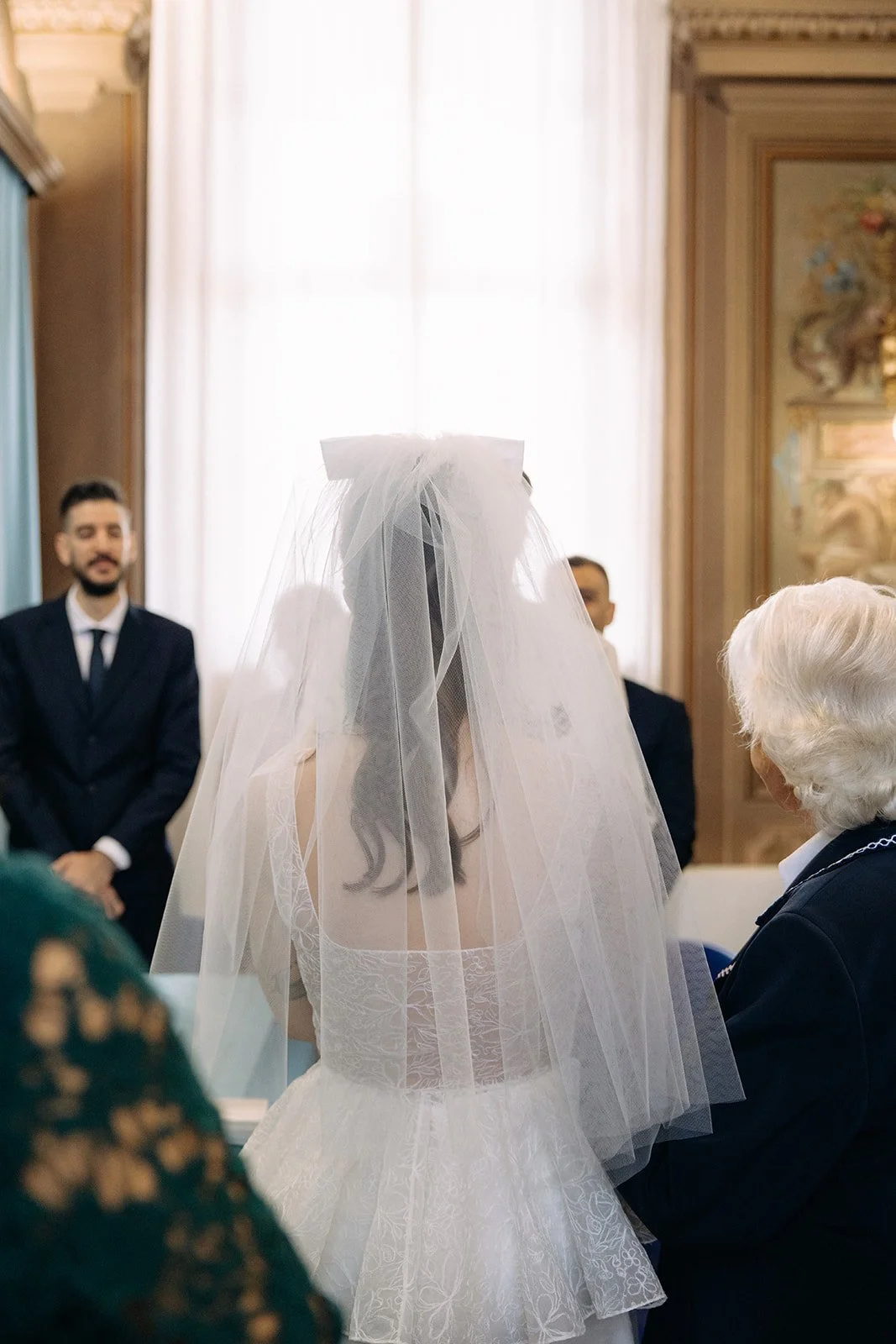Bride wearing a wedding dress and veil during a wedding ceremony, with family members and officiant present in a decorated room.