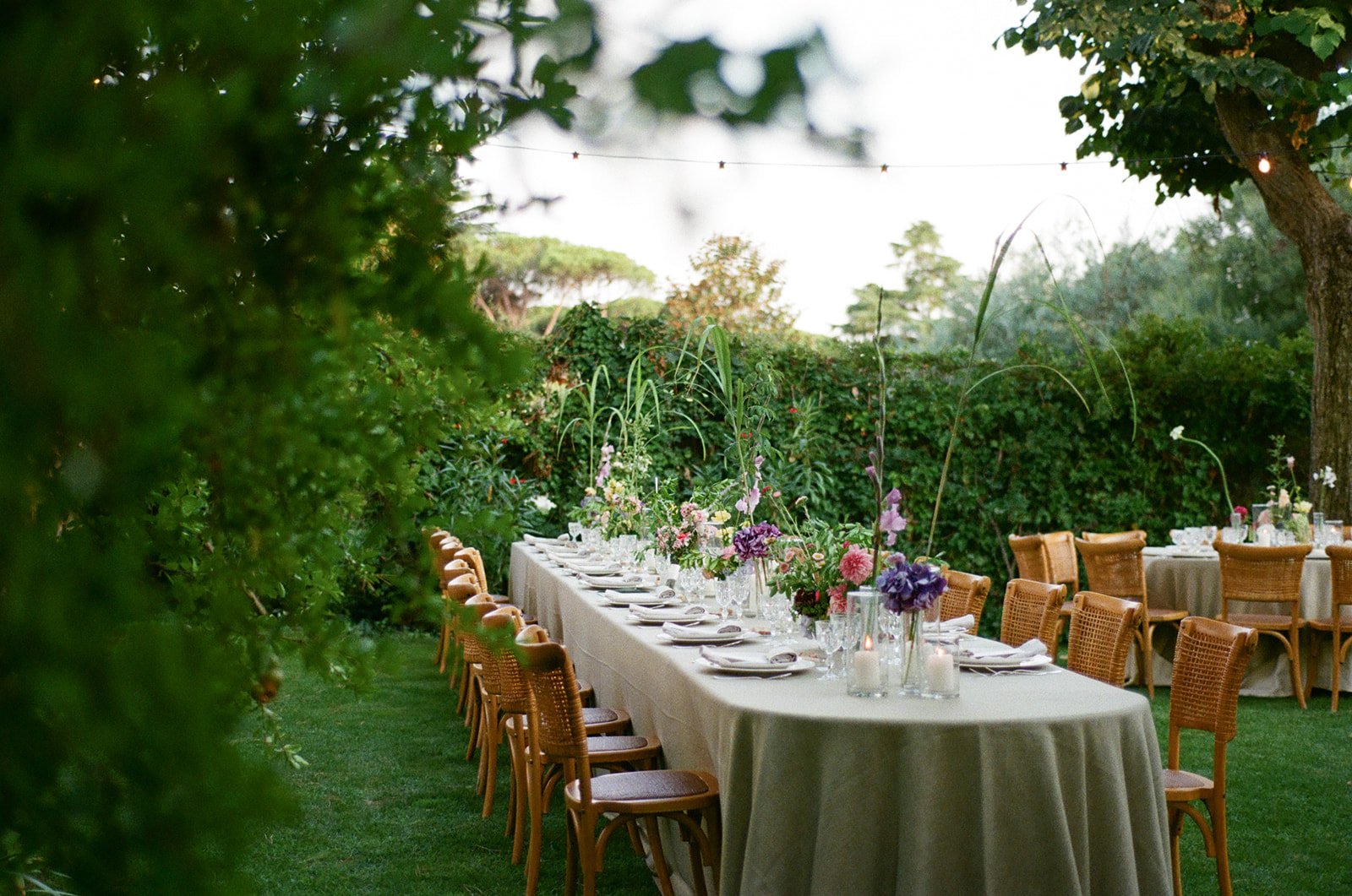 A long outdoor dining table set with white tablecloths, floral centerpieces, and place settings, arranged in a garden with trees, bushes, and string lights overhead.