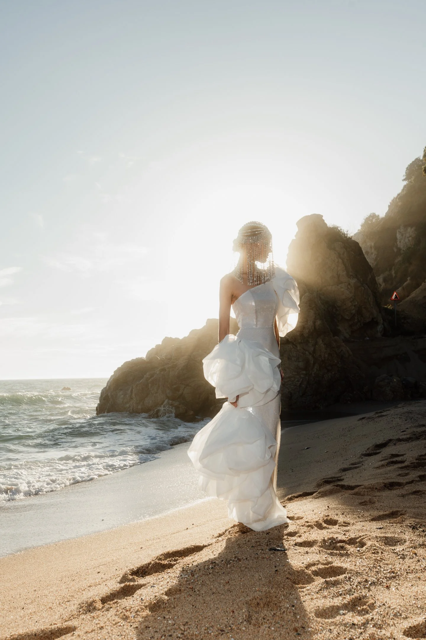 A woman in a white wedding dress standing on a beach at sunset with rocks in the background.