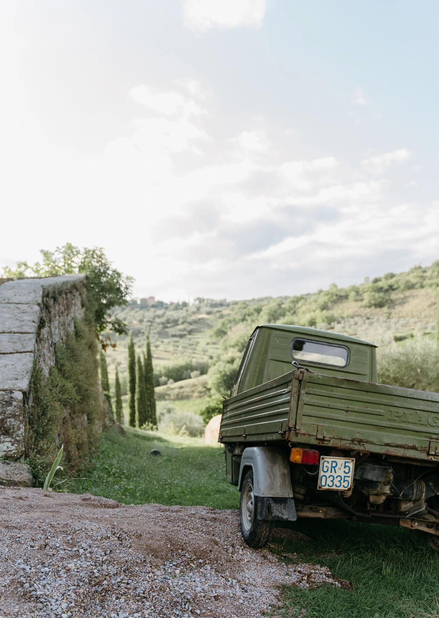 A green pickup truck parked next to a stone wall on a grassy hillside, overlooking rolling hills and sparse trees under a partly cloudy sky.